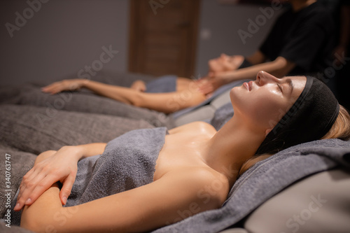 side view on calm relaxed caucasian woman lying in wellbeing salon before skin treatment  woman get face lift massage in the background