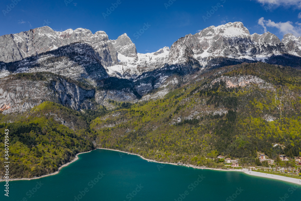 The Improbable aerial landscape of village Molveno, Italy, azure water of lake, empty beach, snow covered mountains Dolomites on background, roof top of chalet, sunny weather, a piers, coastline, 