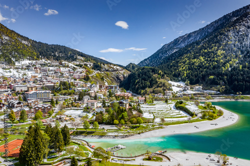 Fototapeta Naklejka Na Ścianę i Meble -  The Improbable aerial landscape of village Molveno, Italy, azure water of lake, empty beach, snow covered mountains Dolomites on background, roof top of chalet, sunny weather, a piers, coastline, 
