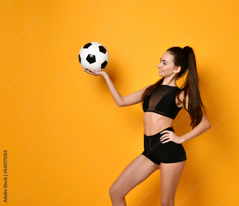 Brunette woman dressed in black top and shorts. Smiling, throw up soccer ball and looking at it, posing on orange background Brunette woman dressed in black top and shorts. Smiling, throw up soccer ball and looking at it, posing on orange background