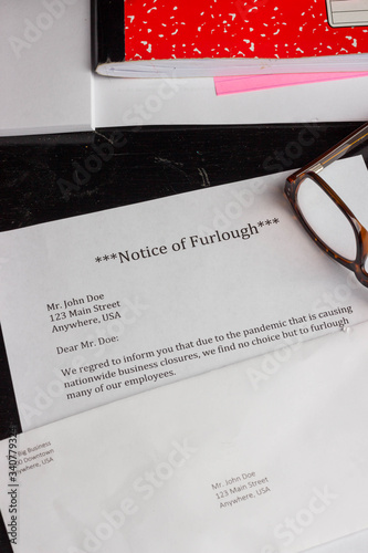 фотографія A notice of furlough lays on a table with an envelope, computer, notebook, and reading glasses