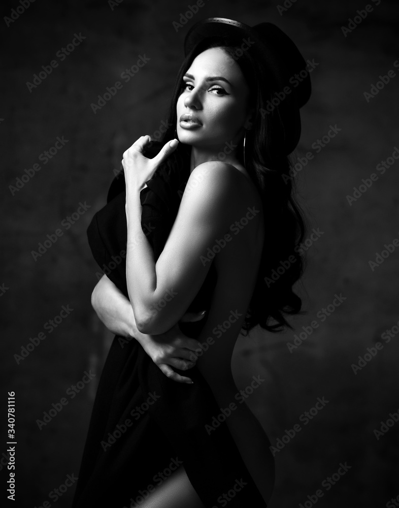 Young beautiful stylish brunette naked woman in hat standing and holding black clothing in hands. Black and white image Young beautiful stylish brunette naked woman in hat standing and holding black clothing in hands. Black and white image