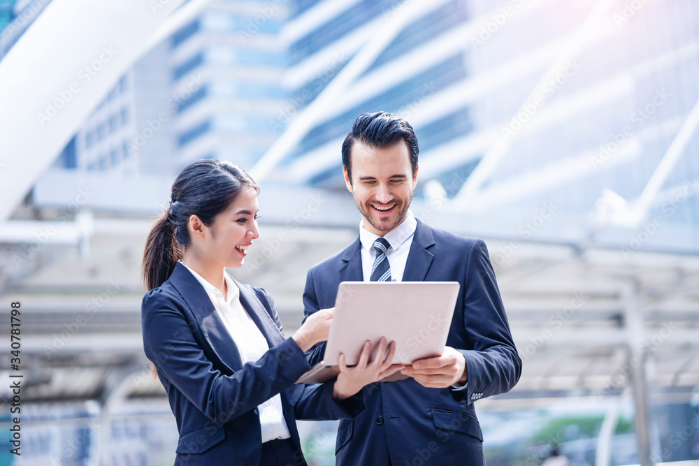 asian businesswoman holding a laptop computer with caucasian businessman showing and explaining business work information, outdoor in the city district with urban architectural structure in background asian businesswoman holding a laptop computer with caucasian businessman showing and explaining business work information, outdoor in the city district with urban architectural structure in background