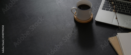 Cropped shot of dark modern office desk with laptop, coffee cup, notebooks and copy space