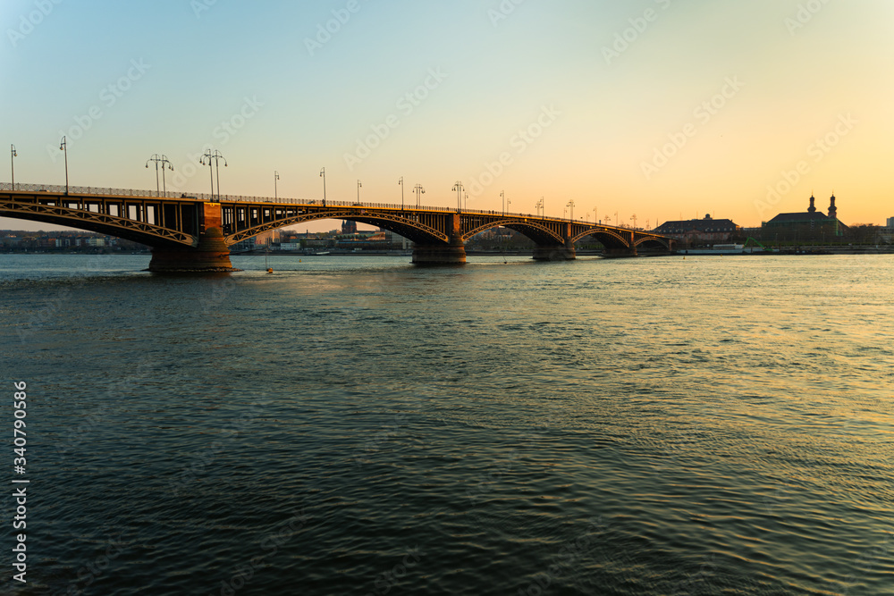 Die Theodor-Heuss-Brücke ragt umgeben vom wunderschön blauen Rhein im Sonnenuntergang in die mainzer Skyline