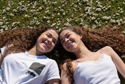 Twin teenage sisters laughing and laying on the grass with daisies on their heads.