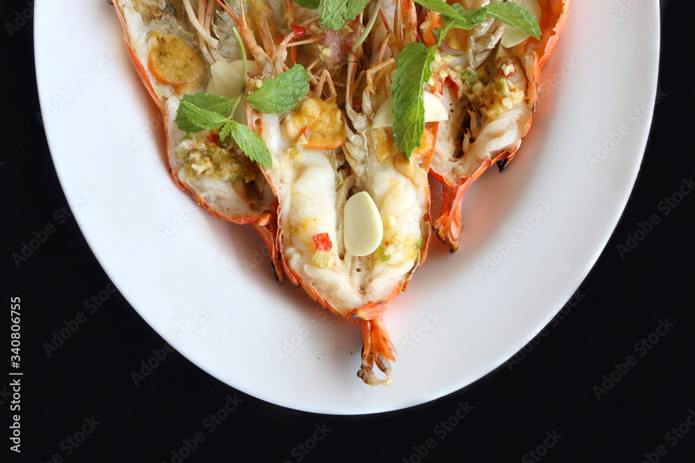 Top view, close up grilled fresh river shrimp with seafood sauce, slice garlic and mint leaves in white plate on black background is famous food in Thailand look delicious.