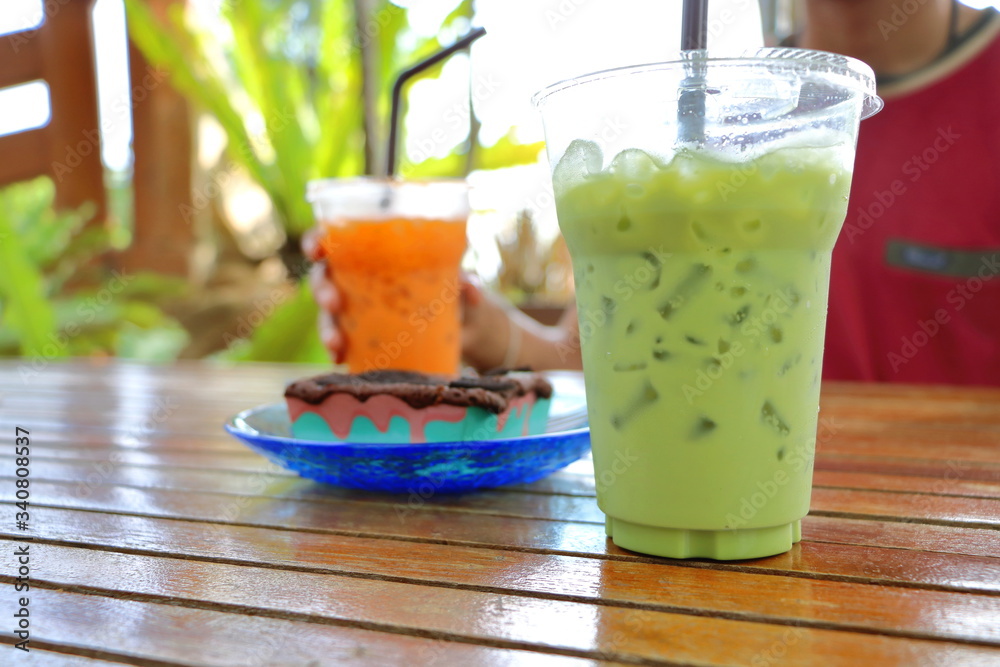 Brownie chocolate cake have cookies, chocolate- chip on topping and have fork beside on blue dish and soft drink (glass of green tea milk and orange Thai tea milk) on wood table, people hold one glass Brownie chocolate cake have cookies, chocolate- chip on topping and have fork beside on blue dish and soft drink (glass of green tea milk and orange Thai tea milk) on wood table, people hold one glass