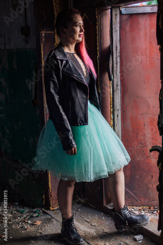 Portrait of a young girl with pink hair standing next to the old rusty door inside of collapsed building surrounded by ruins