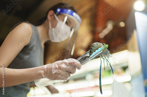 Woman wearing gloves, face shield and mask choosing fresh shrimp at supermarket. Panic shopping during the corona virus pandemic.