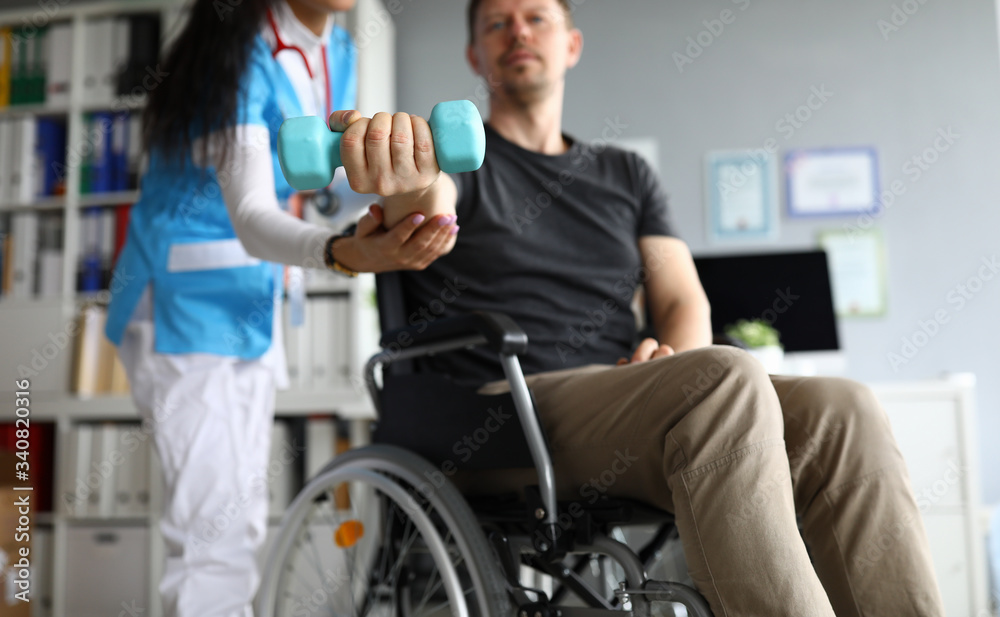 Man on wheelchair holds dumbbell in his hands. Doctor and young man in wheelchair. Treatment and restoration lost functions. Rehabilitation after injuries. Restoration motor functions hand Man on wheelchair holds dumbbell in his hands. Doctor and young man in wheelchair. Treatment and restoration lost functions. Rehabilitation after injuries. Restoration motor functions hand