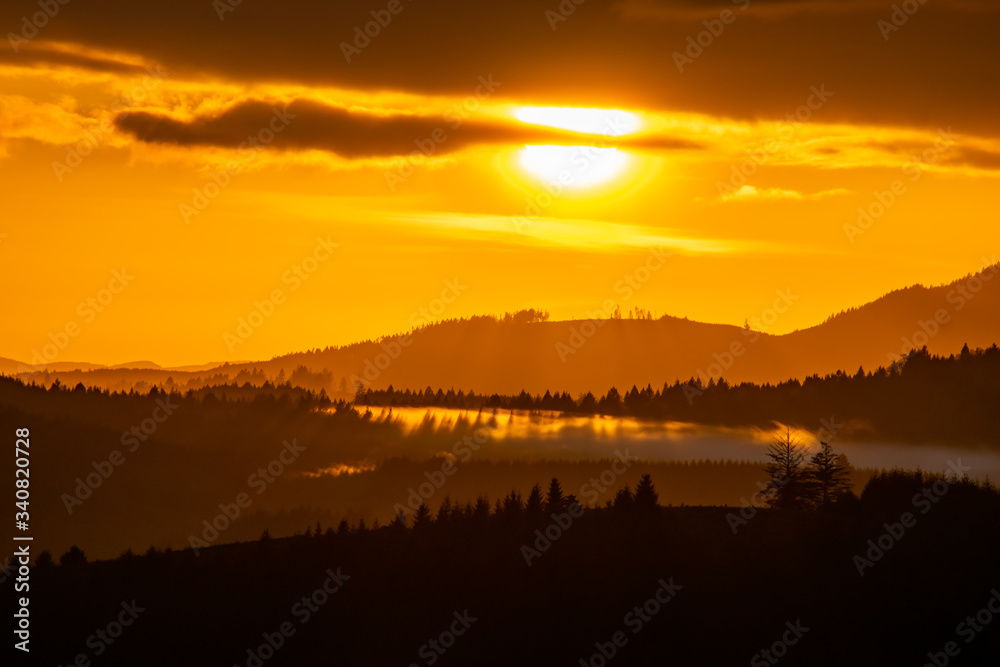 Vivid color bathes distant hills and fog as the sun sets in the distance, edges of trees on hills, shadows on the fog, and streaks of gold in the sky.