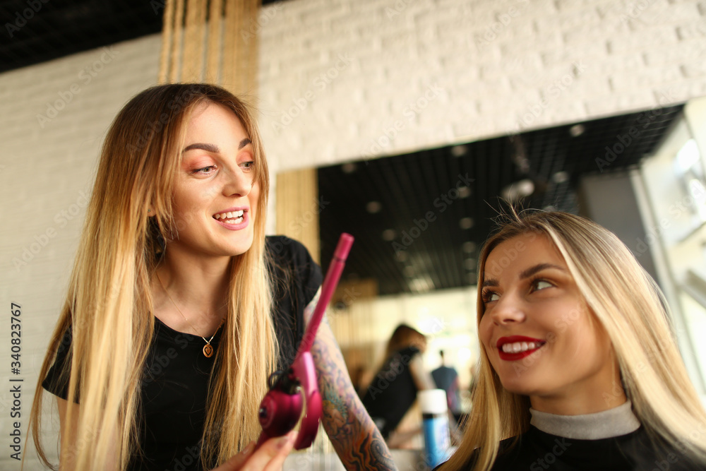 Happy Hairdresser Showing Curling Iron to Client. Blonde Woman Styling Wavy Haircut. Hairstylist Holding Electric Tool for Making Curls. Smiling Young Girl in Beauty Salon for Getting Curly Hairdo
