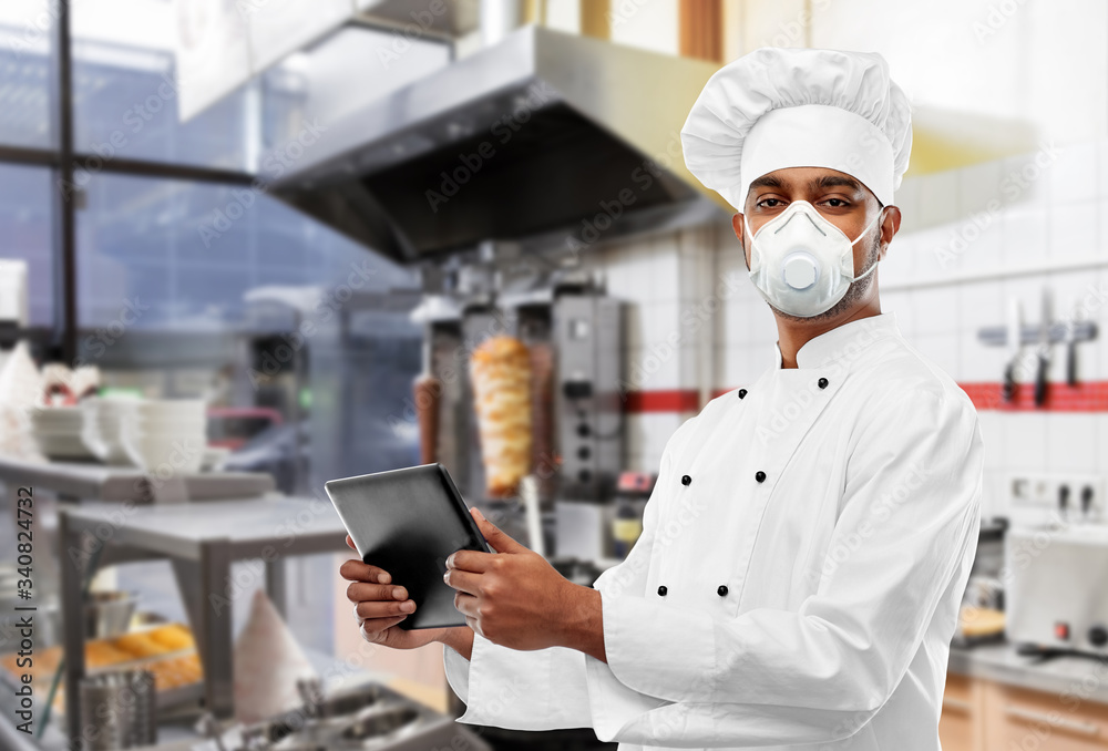 health protection, safety and pandemic concept - male indian chef in toque wearing face protective mask or respirator with tablet computer over kebab shop kitchen background