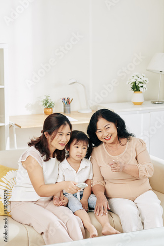 LIttle girl, her mother and grandmother sitting on sofa and watching movie on tv set