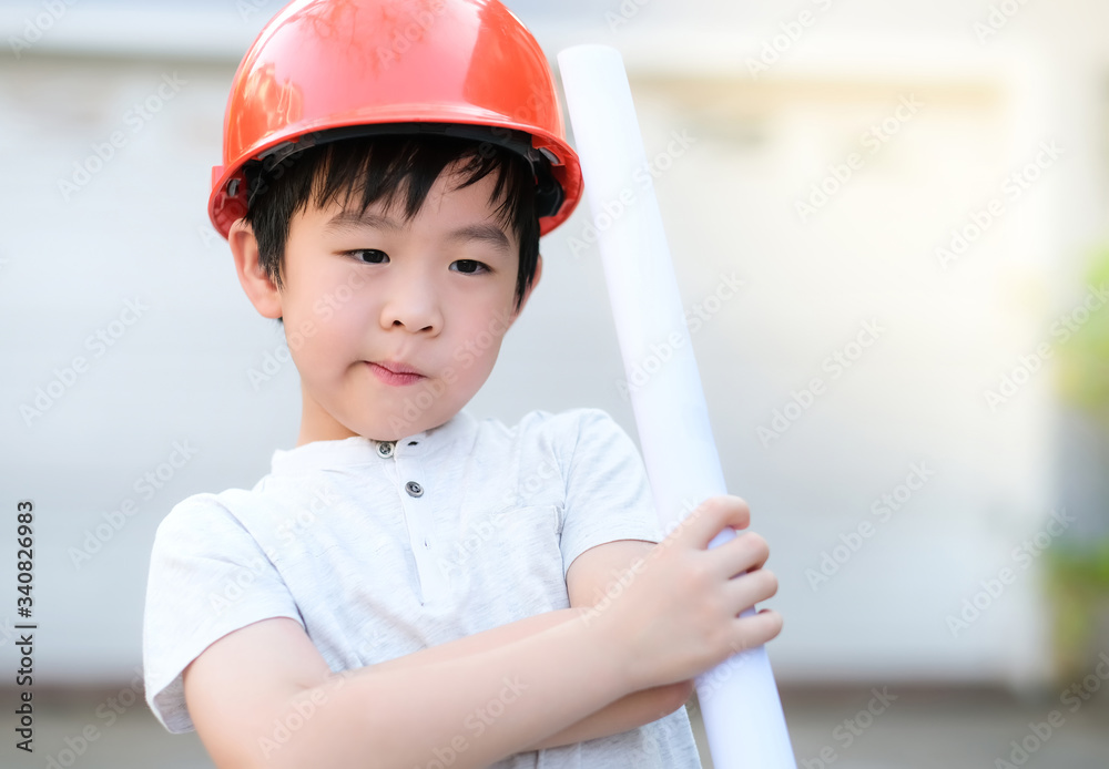 Young Asia boy in gray T shirt wearing a orange safety helmet and holding blueprints in the garden at home under sunlight with blurred background. Engineering and learning education concept. Young Asia boy in gray T shirt wearing a orange safety helmet and holding blueprints in the garden at home under sunlight with blurred background. Engineering and learning education concept.