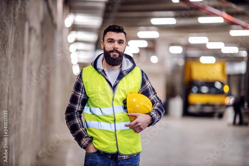Cute bearded construction worker in vest and with safety helmet in hands standing inside of building he renovating.