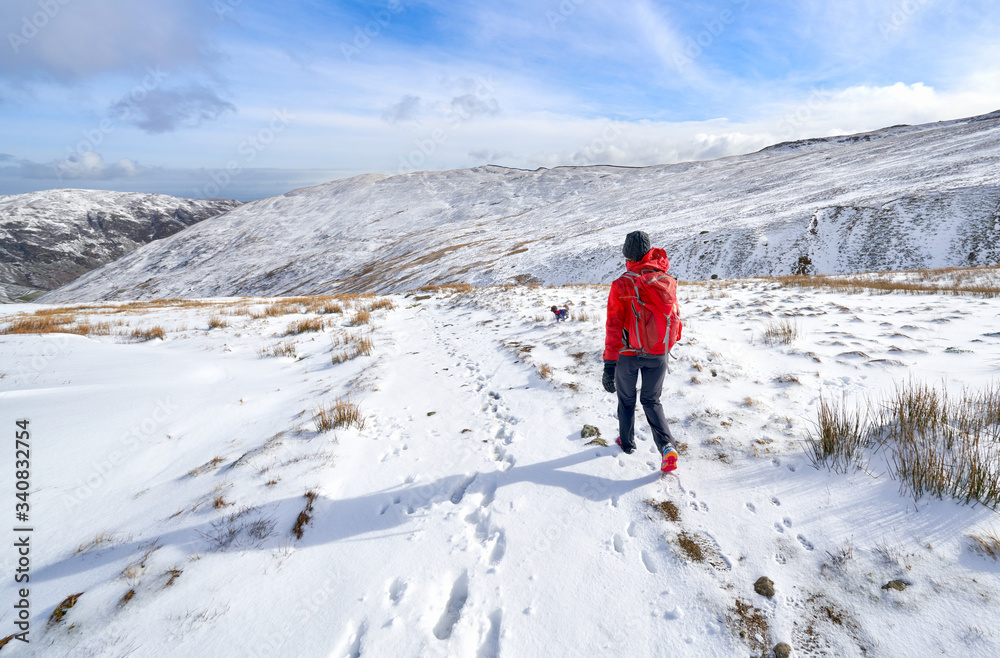 A hiker wearing a bright red jacket walking down from a snow covered Catstye Cam towards Glenridding on a cold sunny winters day in the Lake District, UK.