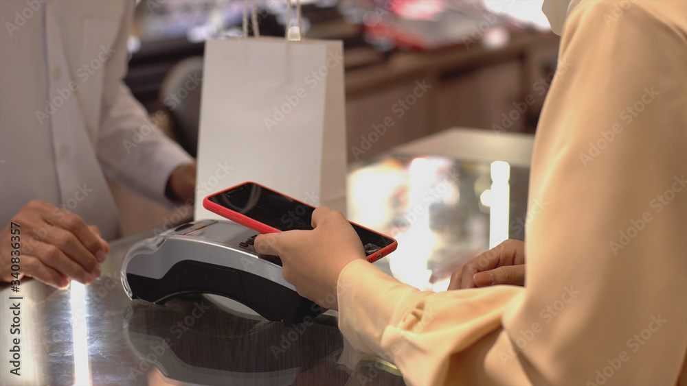 An upwardly mobile Asian Muslim woman using a mobile phone - smartwatch to pay for a product at a sale terminal with nfc identification payment for verification and authentication