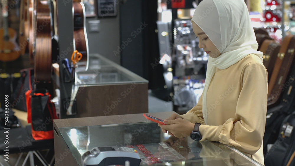 An upwardly mobile Asian Muslim woman using a mobile phone - smartwatch to pay for a product at a sale terminal with nfc identification payment for verification and authentication An upwardly mobile Asian Muslim woman using a mobile phone - smartwatch to pay for a product at a sale terminal with nfc identification payment for verification and authentication