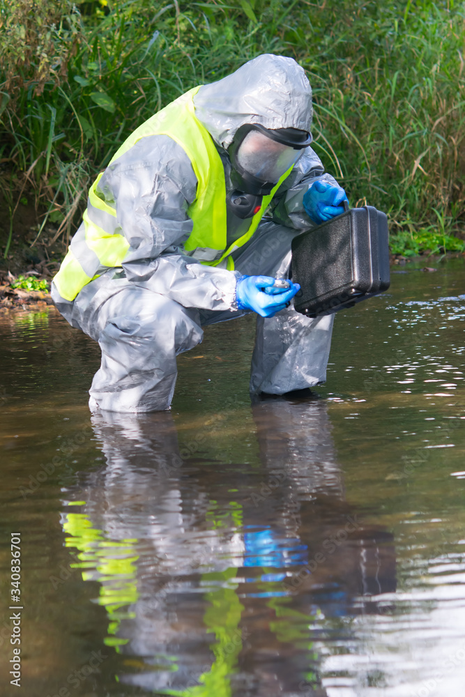 a specialist in a protective suit and mask, with a suitcase, is measuring the device, holding it in his hands, on the pond a specialist in a protective suit and mask, with a suitcase, is measuring the device, holding it in his hands, on the pond