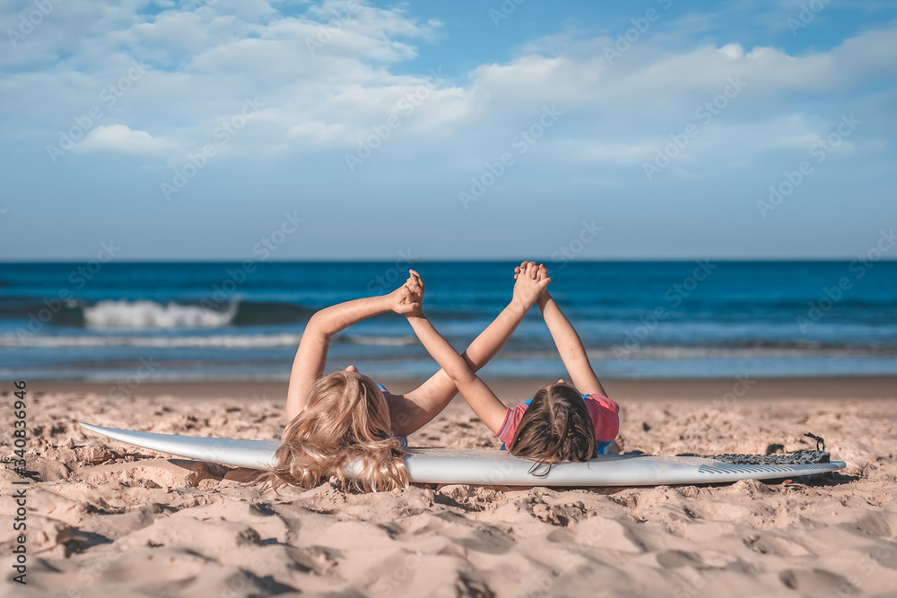 Two little girls' hands holding together laying on a surfboard on the sandy ocean beach. Love, freindship, togetherness concept.