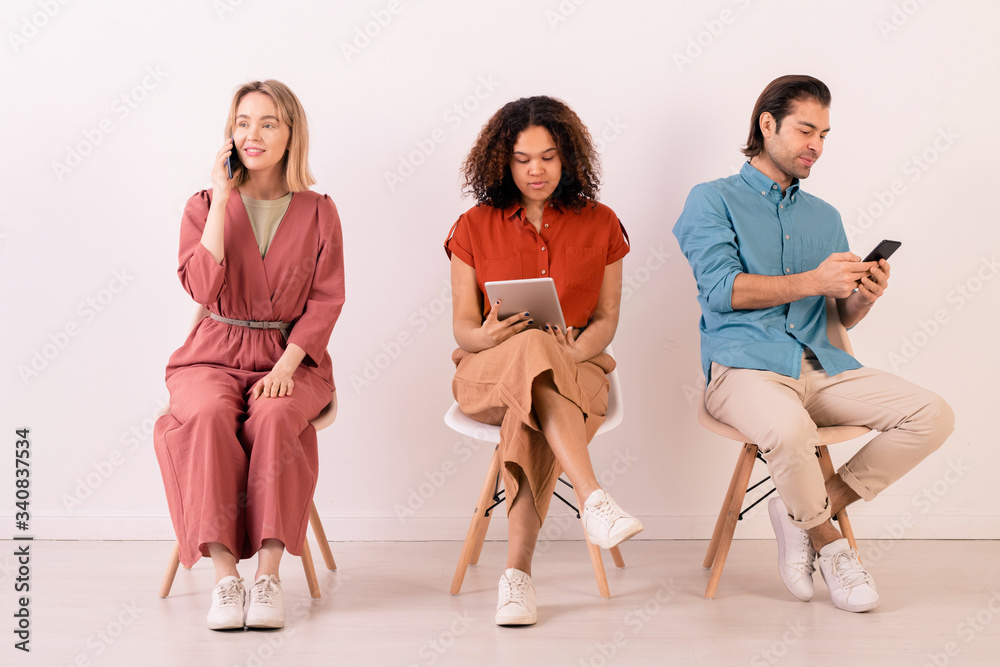 Obraz premium Modern multi-ethnic people in casual outfits sitting on chairs in row and using gadgets all time for communication