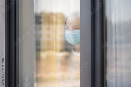 Concept of coronavirus quarantine. Child wearing medical protective face mask during flu virus, looking out of window. COVID-19 - self isolation. Teen girl forced to stay at home. View from street.