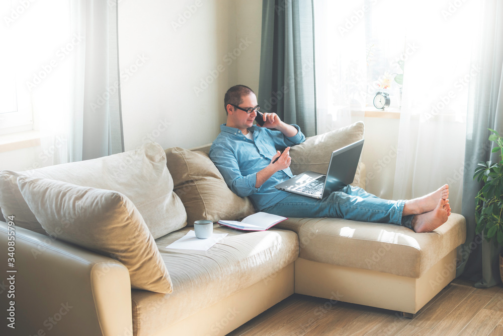 Young man is siiting on the floor and working at home on a computer with the phone, papers and a cup of coffee. Remote work, work from home.