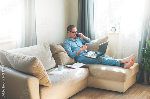 Young man is siiting on the floor and working at home on a computer with the phone, papers and a cup of coffee. Remote work, work from home. © alurk