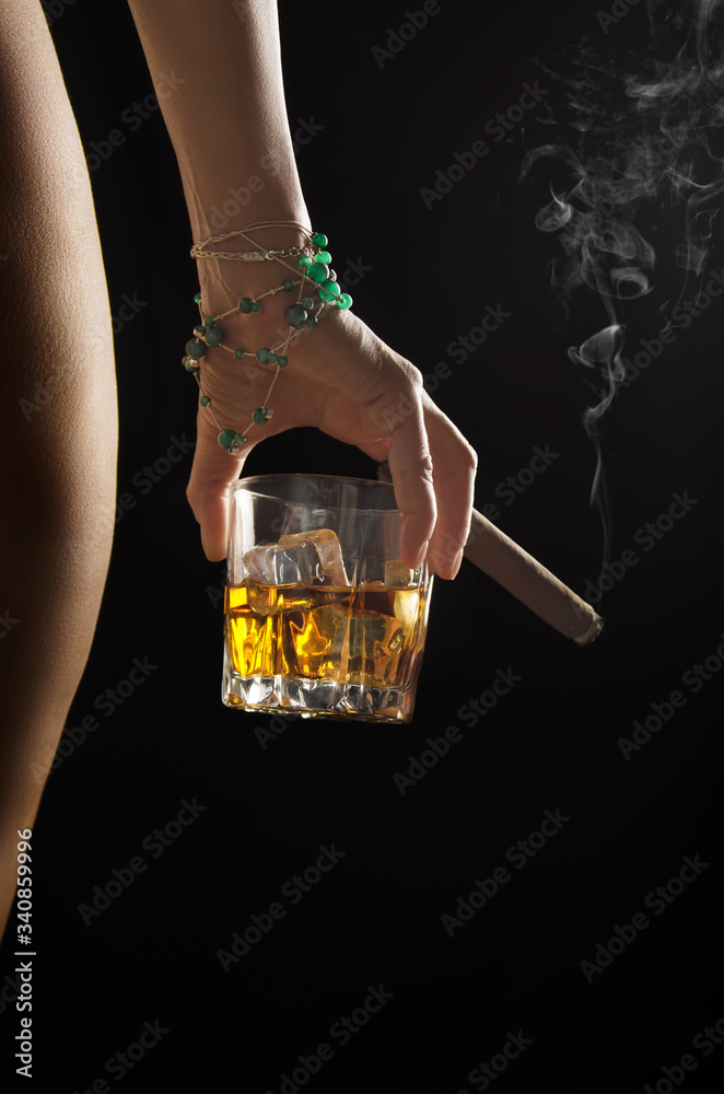 Caucasian model holds glass of whiskey with icecubes and smoking cigar in her hand near naked buttock and hip on dark background closeup studio shot