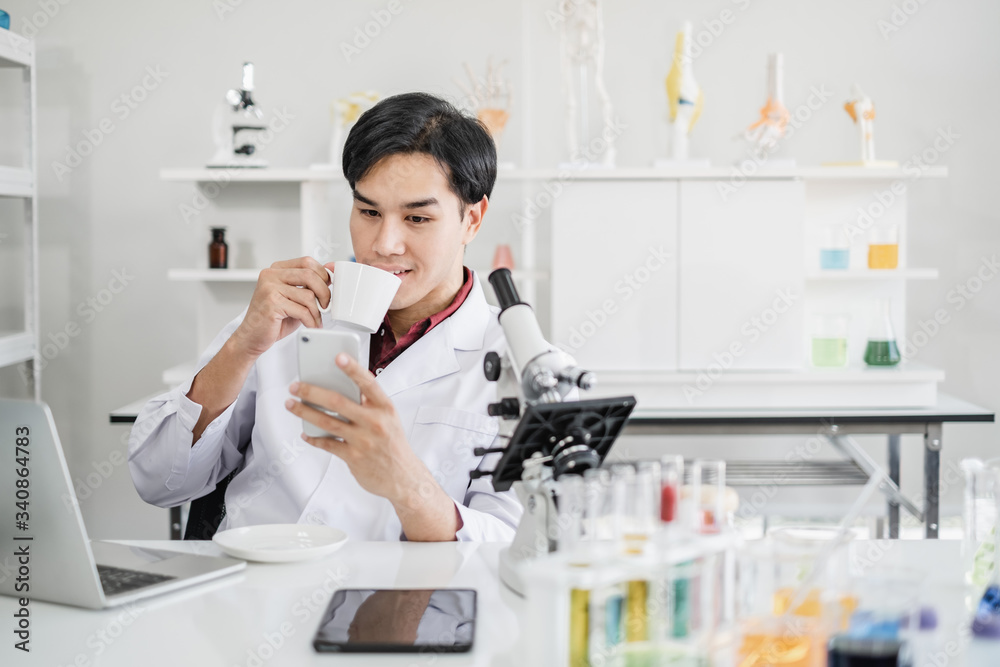 A male scientist with black hair wearing white coat using a mobile phone device sipping coffee with a laptop and a microscope in a laboratory setting with test tubes. Looking relax during break time. A male scientist with black hair wearing white coat using a mobile phone device sipping coffee with a laptop and a microscope in a laboratory setting with test tubes. Looking relax during break time.