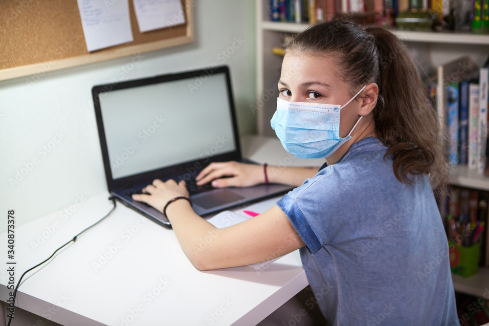 Preteen Caucasian girl in medical mask using her laptop, printing on keyboard, schoolchild studying at home during covid-19 pandemic