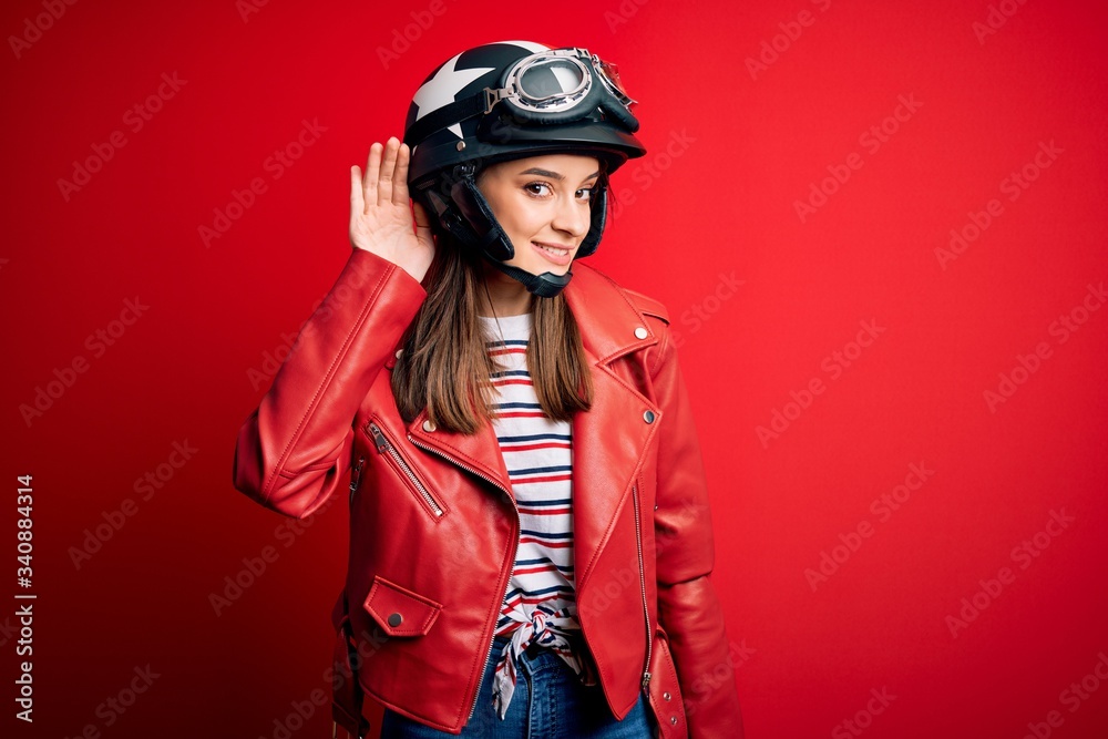 Young beautiful brunette motocyclist woman wearing motorcycle helmet and red jacket smiling with hand over ear listening an hearing to rumor or gossip. Deafness concept.