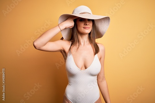 Young beautiful brunette woman on vacation wearing swimsuit and summer hat pointing unhappy to pimple on forehead, ugly infection of blackhead. Acne and skin problem