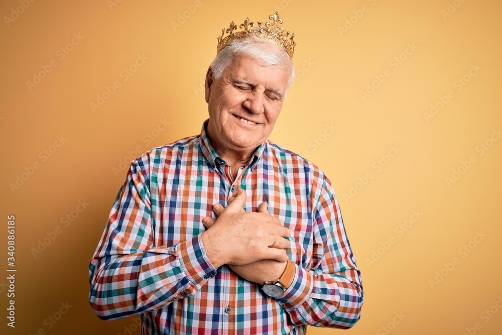 Senior handsome hoary man wearing golden crown of king over isolated yellow background smiling with hands on chest with closed eyes and grateful gesture on face. Health concept.