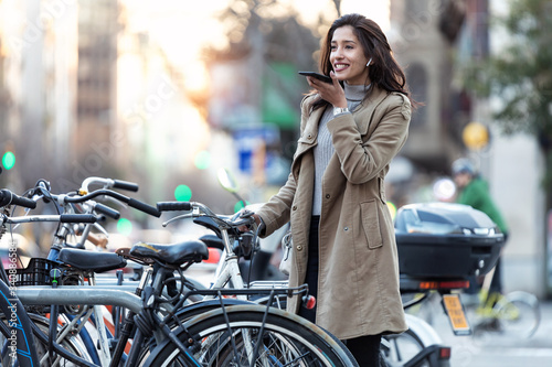 Cuadro en lienzo Pretty young woman using voice recognition system on her smartphone while standing in the street