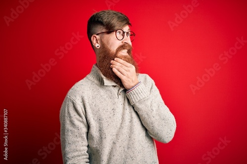 Handsome Irish redhead man with beard wearing casual sweater and glasses over red background Thinking worried about a question, concerned and nervous with hand on chin © Krakenimages.com