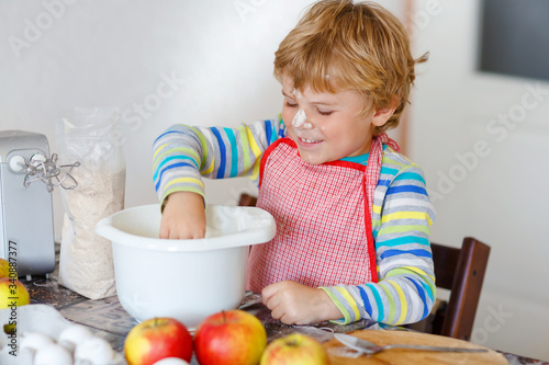 Cute little happy blond preschool kid boy baking apple cake and muffins in domestic kitchen. Funny lovely healthy child having fun with working with mixer, flour, eggs, fruits. Little helper indoors