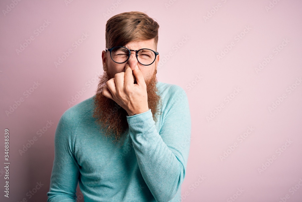 Handsome Irish redhead man with beard wearing glasses over pink isolated background smelling something stinky and disgusting, intolerable smell, holding breath with fingers on nose. Bad smell Handsome Irish redhead man with beard wearing glasses over pink isolated background smelling something stinky and disgusting, intolerable smell, holding breath with fingers on nose. Bad smell