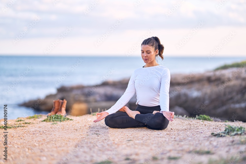 Young beautiful sportwoman smiling happy practicing yoga. Coach with smile on face teaching lotus pose at the beach