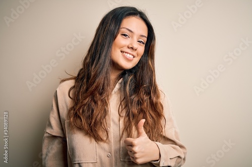 Young beautiful brunette woman wearing casual shirt standing over white background doing happy thumbs up gesture with hand. Approving expression looking at the camera showing success.