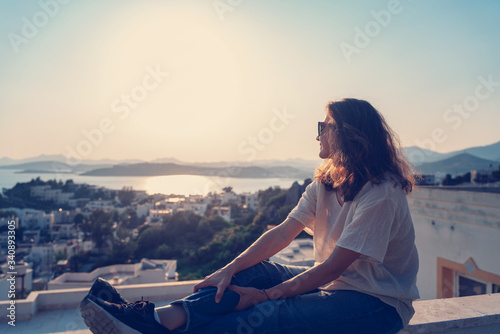 Beautiful charming happy young girl woman in sunglasses looks from the balcony at the city the sea and nature at sunset