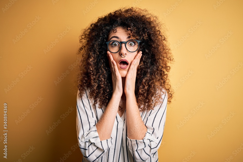 Young beautiful woman with curly hair and piercing wearing striped shirt and glasses afraid and shocked, surprise and amazed expression with hands on face Young beautiful woman with curly hair and piercing wearing striped shirt and glasses afraid and shocked, surprise and amazed expression with hands on face