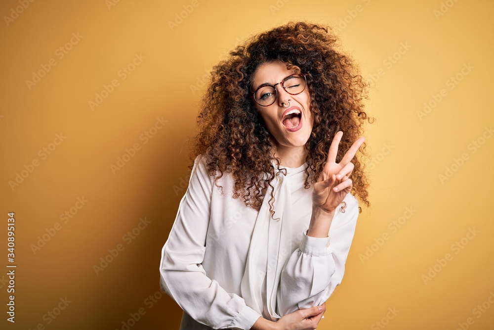 Young beautiful brunette woman with curly hair and piercing wearing shirt and glasses smiling with happy face winking at the camera doing victory sign. Number two. Young beautiful brunette woman with curly hair and piercing wearing shirt and glasses smiling with happy face winking at the camera doing victory sign. Number two.