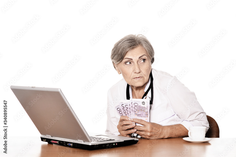 Senior doctor sitting at table with laptop and holding money Senior doctor sitting at table with laptop and holding money