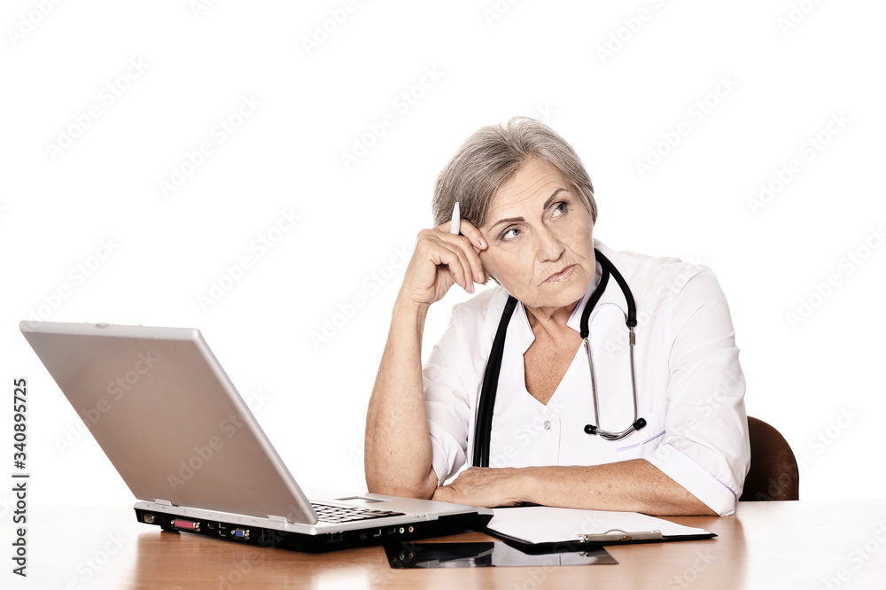 Serious elderly woman doctor sitting at table with computer on white Serious elderly woman doctor sitting at table with computer on white