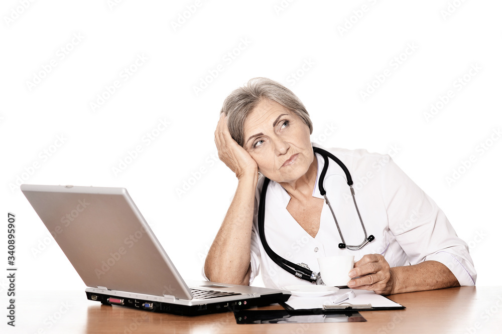 Serious elderly woman doctor sitting at table with computer on white Serious elderly woman doctor sitting at table with computer on white