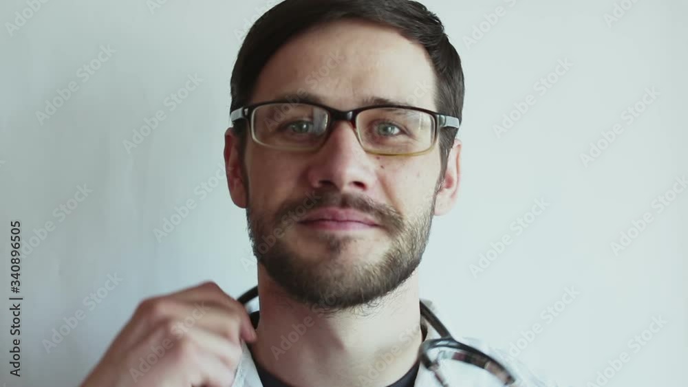 A young handsome doctor wearing glasses smiles broadly and takes off a stethoscope and hangs it on his neck. A young doctor is holding a stethoscope. A young handsome doctor wearing glasses smiles broadly and takes off a stethoscope and hangs it on his neck. A young doctor is holding a stethoscope.