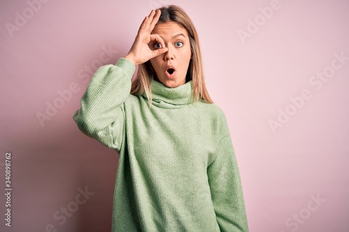 Young beautiful blonde woman wearing winter wool sweater over pink isolated background doing ok gesture shocked with surprised face, eye looking through fingers. Unbelieving expression. © Krakenimages.com